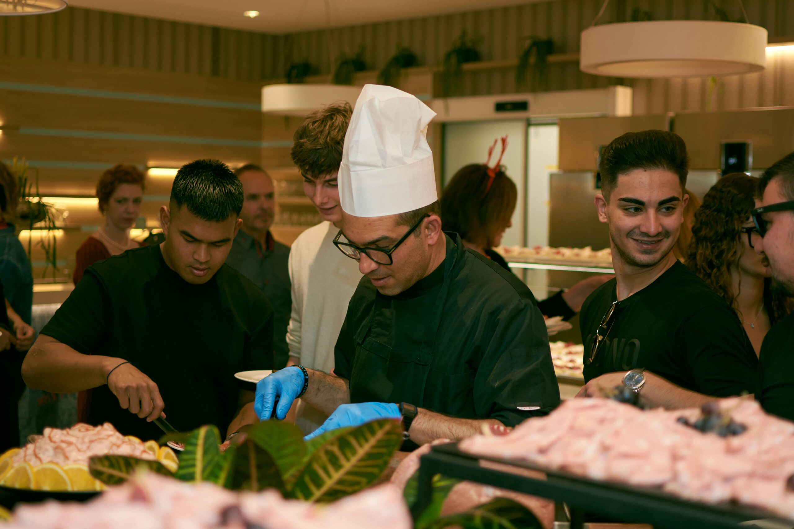A chef in glasses serves food to guests at a lively indoor buffet event.