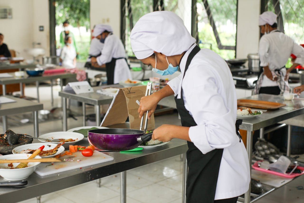 Culinary students in uniforms practice cooking skills in a professional kitchen setting.